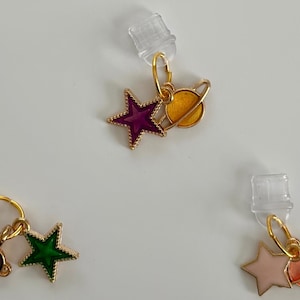 May include: Gold-toned earrings featuring celestial designs. Each earring has a star and planet charm with colorful enamel details. The stars are purple, green, and pink. The planets are yellow, maroon, and orange. The earrings have clear earring backs.