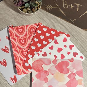 Puede incluir: Una colección de tarjetas con temática de corazones en varios diseños y colores. Las tarjetas presentan patrones de corazones rojos, corazones de acuarela y contornos de corazones. En el fondo se ve una pequeña planta suculenta en una maceta blanca.