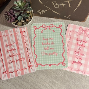 May include: Three decorative cards with the text "buy me books & tell me I'm pretty". The cards have pink and green accents, with bow and gingham patterns. A small succulent plant in a white pot is in the background.
