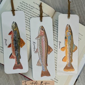 May include: Three rectangular white bookmarks featuring illustrations of fish labelled Brook, Rainbow, and Brown. Each bookmark has a hole at the top with a brown leather cord tied through it. The fish illustrations are detailed with colours including orange, brown, and pink.