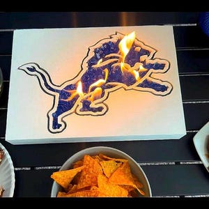 May include: A white fire pit with a blue Detroit Lions logo. The fire pit is lit and has flames coming out of the logo. There are snacks and drinks on the table around the fire pit.