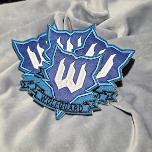 May include: A cluster of embroidered patches featuring a blue and white design. Each patch has a shield shape with a stylized "W" and the text "WOLFGURAD" on a banner. The patches are on a gray fabric.