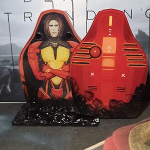 May include: Two red and black decorative figures. One figure features a portrait of a person with long brown hair, wearing a red and yellow outfit. The other figure is red with orange and black details. Both figures are on a black base.