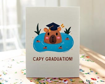 May include: A graduation card featuring a cartoon capybara wearing a graduation cap and floating in a pool of water. The card has the text "Capy Graduation!" printed on it.