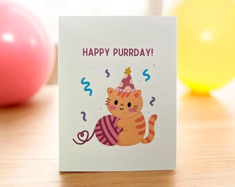 May include: A white card with the text "Happy Purrday!" and a cartoon cat wearing a party hat and holding a ball of yarn. The cat is orange and white with a pink nose and whiskers.