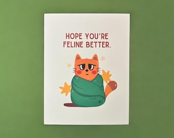 May include: A greeting card with a cartoon orange cat wrapped in a green blanket. The card says "Hope you're feline better."