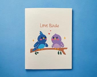 May include: A greeting card with a light pink background featuring two cartoon birds on a branch. The blue bird is on the left and the purple bird is on the right. The text "Love Birds" is printed in orange at the top of the card.