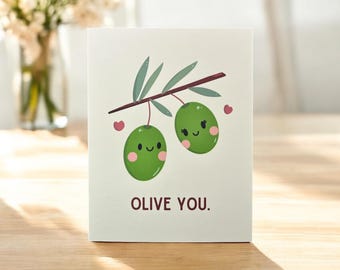 May include: A greeting card with a green olive branch with two olives with faces on them. The olives are smiling and have hearts around them. The text on the card says "Olive You."