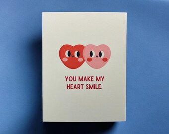 May include: A white card with two red and pink hearts with smiling faces. The text on the card reads "You make my heart smile."