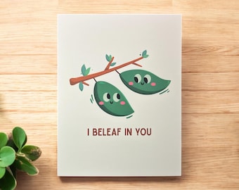 May include: A greeting card with a whimsical illustration of two green leaves with smiling faces hanging from a brown branch. The text "I BELEAF IN YOU" is printed below the leaves.