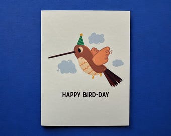 May include: A greeting card with a cartoon hummingbird wearing a party hat, flying among blue cloud illustrations. The text "HAPPY BIRD-DAY" is printed below the bird. The card is on a blue background.