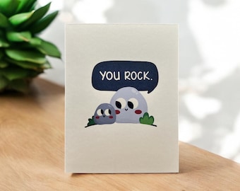 May include: A greeting card with a blue speech bubble that says "You Rock."  Two cartoon rocks with eyes and smiles are in front of the speech bubble.