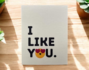 May include: A white card with black text that reads "I like you."  A yellow emoji with red heart eyes is next to the word "you."