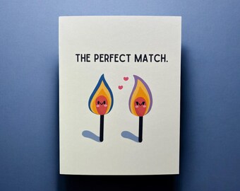 May include: A white card with the text "THE PERFECT MATCH." in black. Two cartoon matchsticks with faces are drawn on the card. The matchsticks are facing each other and have hearts drawn between them.