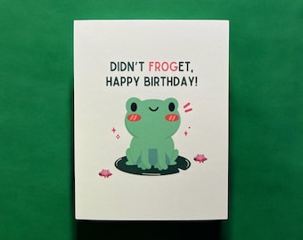 May include: A birthday card with a green frog illustration sitting on a lily pad. The card reads "Didn't Froget, Happy Birthday!" in black and red text. Pink accents and sparkles surround the frog.