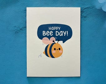 May include: A white card with a blue speech bubble that says "Happy Bee Day!" and a cartoon bee with black stripes and a yellow body.