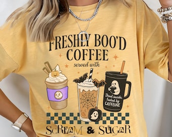 Può includere: T-shirt giallo senape con un design a tema Halloween. La grafica include il testo "FRESHLY BOO'D COFFEE" e illustrazioni di bevande al caffè con elementi spettrali come pipistrelli, zucche e fantasmi. È presente anche la frase "SCREAM & SUGAR".
