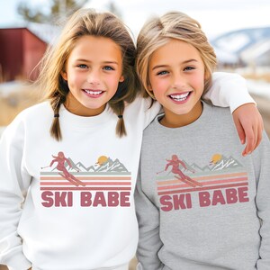 May include: Two children wearing matching sweatshirts with a retro-style graphic of a skier in the mountains and the text "SKI BABE". The sweatshirt on the left is white and the sweatshirt on the right is gray.