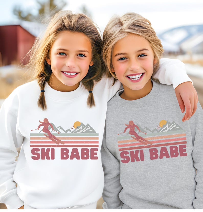 May include: Two children wearing matching sweatshirts with a retro-style graphic of a skier in the mountains and the text "SKI BABE". The sweatshirt on the left is white and the sweatshirt on the right is gray.