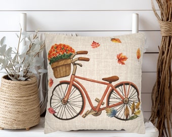 May include: A square throw pillow with a light beige linen-textured cover featuring a watercolor illustration of a red bicycle. The bicycle has a brown wicker basket filled with orange flowers, and several colorful autumn leaves float around it.