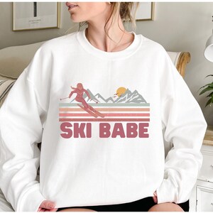 May include: White sweatshirt with a pink graphic of a skier in a mountain landscape with a sunset. The text "SKI BABE" is printed in pink below the graphic.