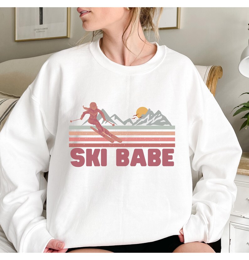 May include: White sweatshirt with a pink graphic of a skier in a mountain landscape with a sunset. The text "SKI BABE" is printed in pink below the graphic.