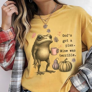 May include: A mustard yellow t-shirt featuring a graphic of a frog holding a pink mug, accompanied by pumpkins and autumn leaves. The text reads, God's got a plan. Mine was terrible. The shirt is worn with a red, white, and grey plaid flannel shirt.