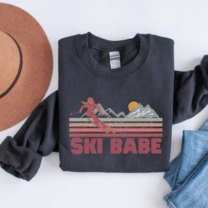 May include: A black sweatshirt with a retro-style graphic of a woman skiing down a mountain with the text "SKI BABE" in red.