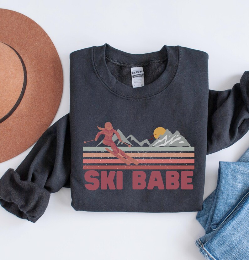 May include: A black sweatshirt with a retro-style graphic of a woman skiing down a mountain with the text "SKI BABE" in red.
