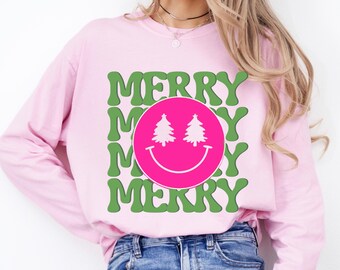May include: A light pink long-sleeve sweatshirt with the word "MERRY" in green, repeated three times. A pink circle with a smiley face and two green Christmas trees is in the center.