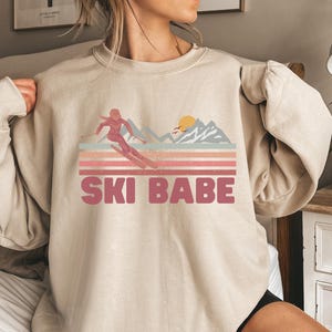 May include: A beige sweatshirt with a pink graphic of a woman skiing down a mountain with a sunset in the background. The text "SKI BABE" is printed below the graphic.