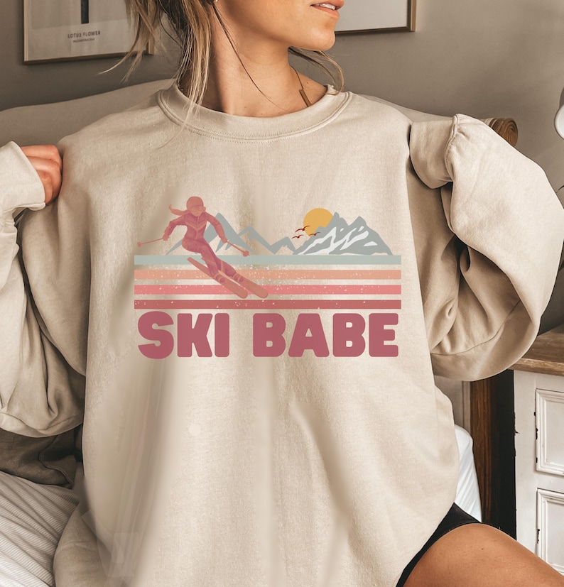 May include: A beige sweatshirt with a pink graphic of a woman skiing down a mountain with a sunset in the background. The text "SKI BABE" is printed below the graphic.