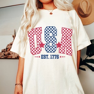 May include: A white t-shirt with a red, white, and blue checkered "USA" graphic and the text "EST. 1776".