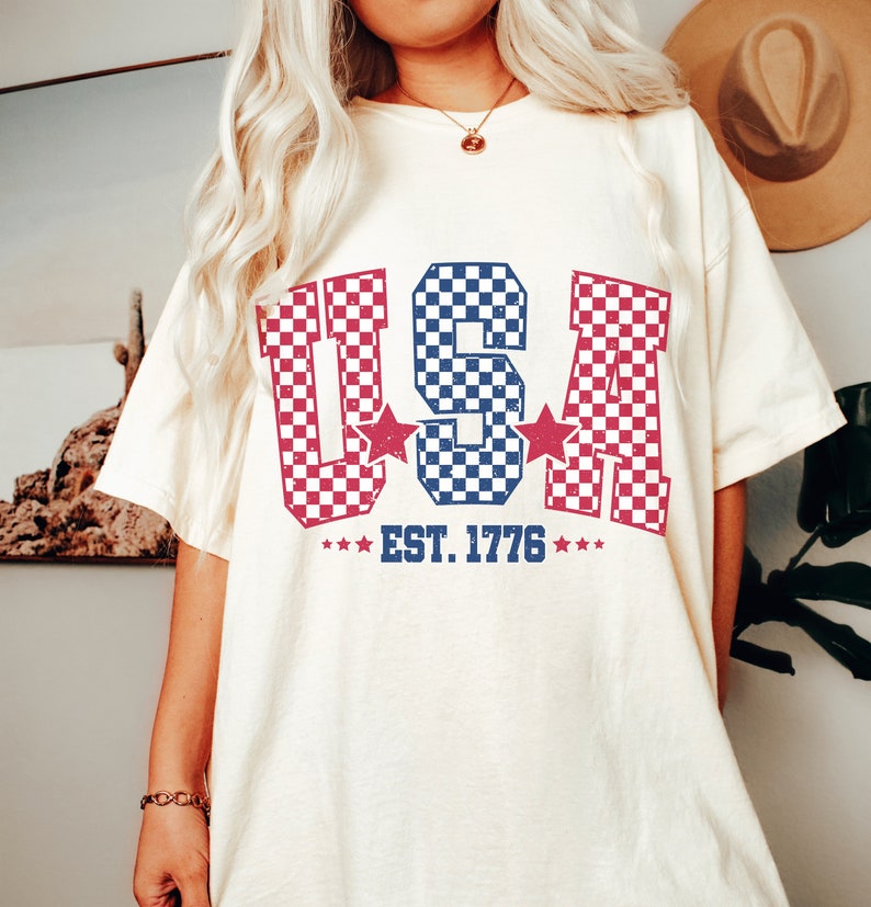 May include: A white t-shirt with a red, white, and blue checkered "USA" graphic and the text "EST. 1776".