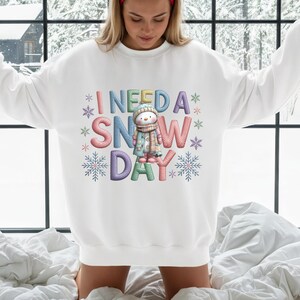 May include: White sweatshirt with the phrase "I NEED A SNOW DAY" in colorful, embroidered letters. A cartoon snowman wearing a hat and scarf is in the center. Snowflakes and stars surround the text, creating a winter theme.