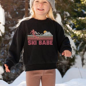 May include: A black sweatshirt with a retro-style graphic of a skier in front of mountains and the text "SKI BABE".