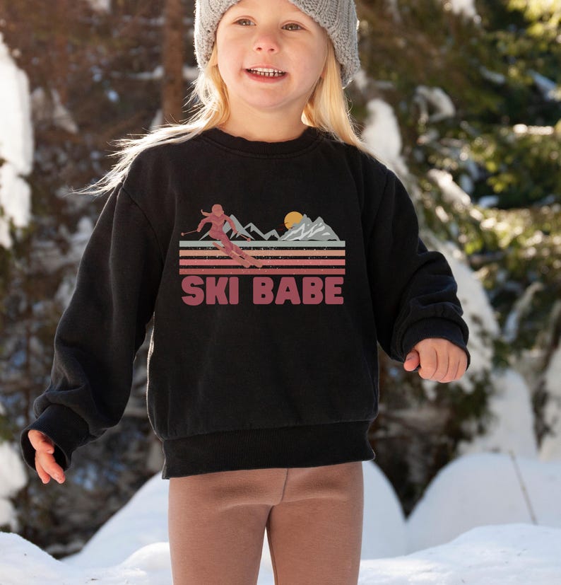May include: A black sweatshirt with a retro-style graphic of a skier in front of mountains and the text "SKI BABE".