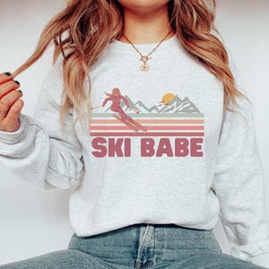 May include: A gray sweatshirt with a retro-style graphic of a woman skiing down a mountain with a sunset in the background. The text "SKI BABE" is printed in pink below the graphic.