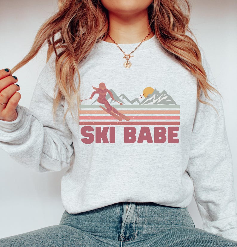 May include: A gray sweatshirt with a retro-style graphic of a woman skiing down a mountain with a sunset in the background. The text "SKI BABE" is printed in pink below the graphic.