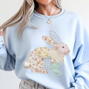 May include: A pale blue sweatshirt with a patchwork rabbit design. The rabbit is crafted from assorted floral and patterned fabrics in pastel shades, including pink, yellow, and blue. The sweatshirt has a classic crew neck and long sleeves.