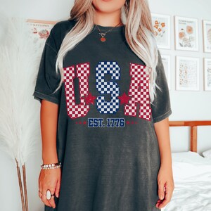 May include: A dark gray t-shirt with a checkered design in red, white, and blue, spelling out "USA" with stars on either side. The text "EST. 1776" is printed below the design.
