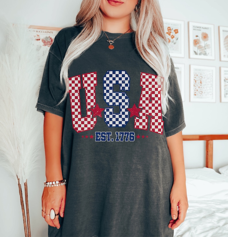 May include: A dark gray t-shirt with a checkered design in red, white, and blue, spelling out "USA" with stars on either side. The text "EST. 1776" is printed below the design.