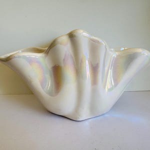 May include: A white ceramic bowl shaped like a seashell, with a pearlescent finish. The bowl has a fluted rim and a textured surface. The iridescent sheen reflects colours, adding a touch of elegance. Suitable for coastal decor, this piece is a decorative item.