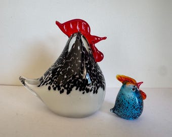 May include: Two glass rooster figurines. The larger rooster is white with black speckles and a red comb. The smaller rooster is blue with orange and red accents. Both are decorative and likely for display.