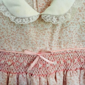 May include: A pink and white dress with a white lace collar and a pink satin ribbon bow. The dress has a smocked bodice and a gathered skirt.