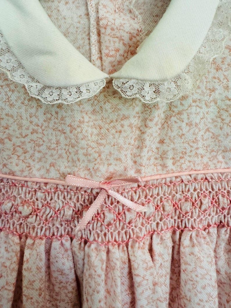May include: A pink and white dress with a white lace collar and a pink satin ribbon bow. The dress has a smocked bodice and a gathered skirt.