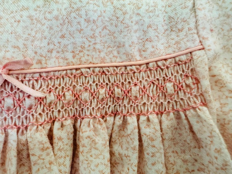 May include: A pink baby dress with a white floral pattern and a pink smocked detail around the waist.
