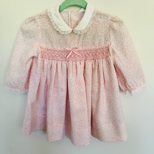 May include: A pink and white floral print dress with a white lace collar and a smocked bodice. The dress has long sleeves and a bow at the waist.