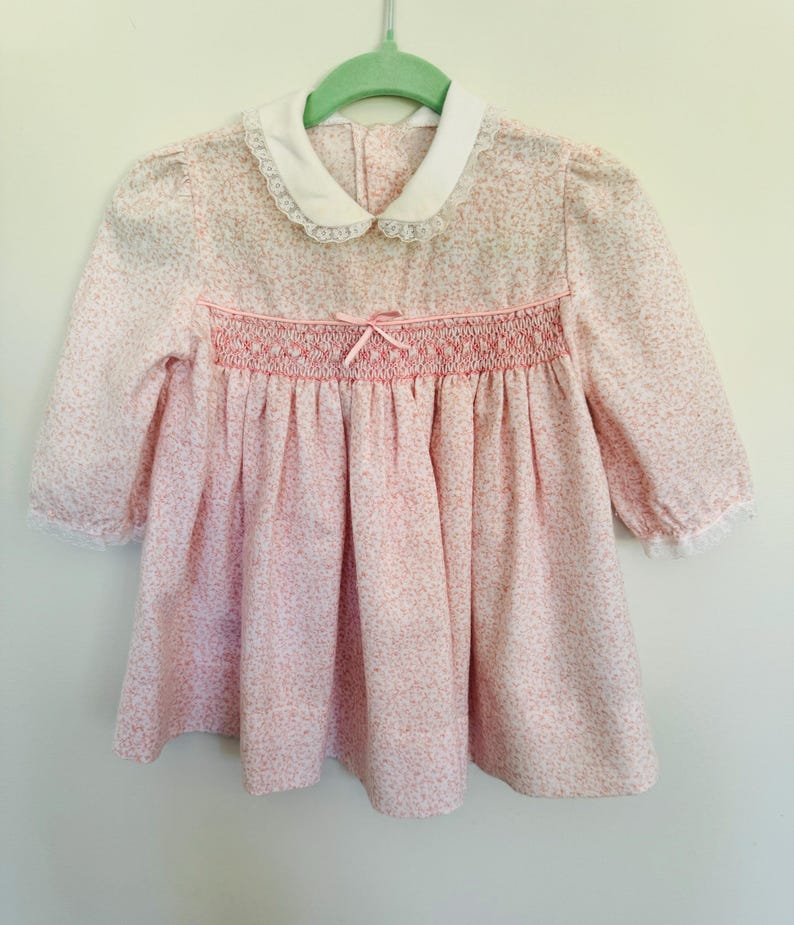 May include: A pink and white floral print dress with a white lace collar and a smocked bodice. The dress has long sleeves and a bow at the waist.