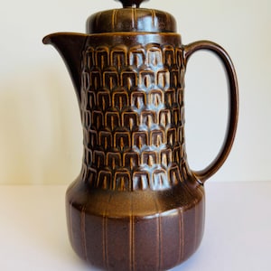May include: A brown ceramic coffee pot with a lid and handle. The body of the pot has a textured, geometric pattern, while the base features vertical lines. The spout is curved, and the handle is a simple loop. The pot is a warm, earthy brown colour.
